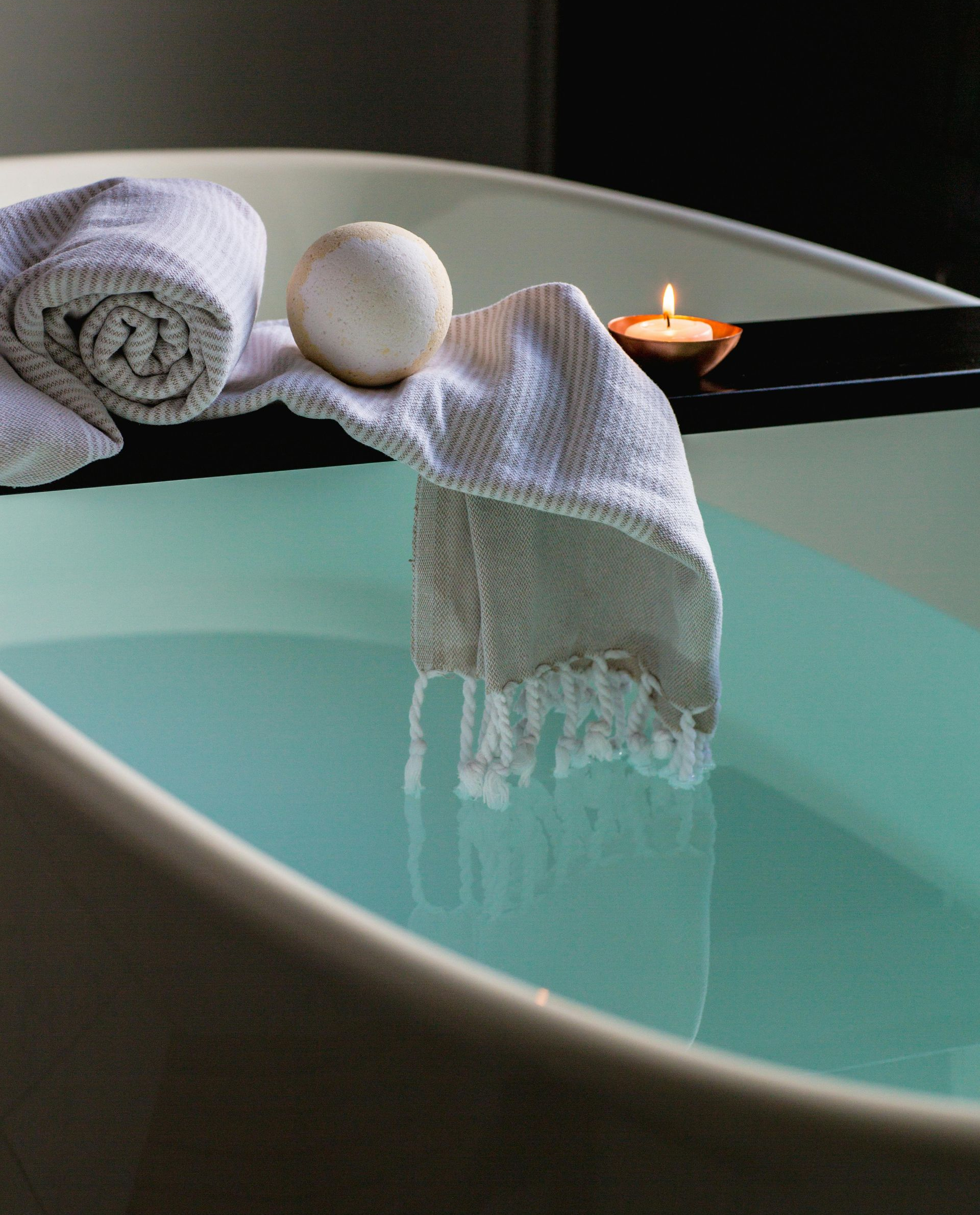 Bathtub filled with blue water, towels, bath bomb, and a lit candle.