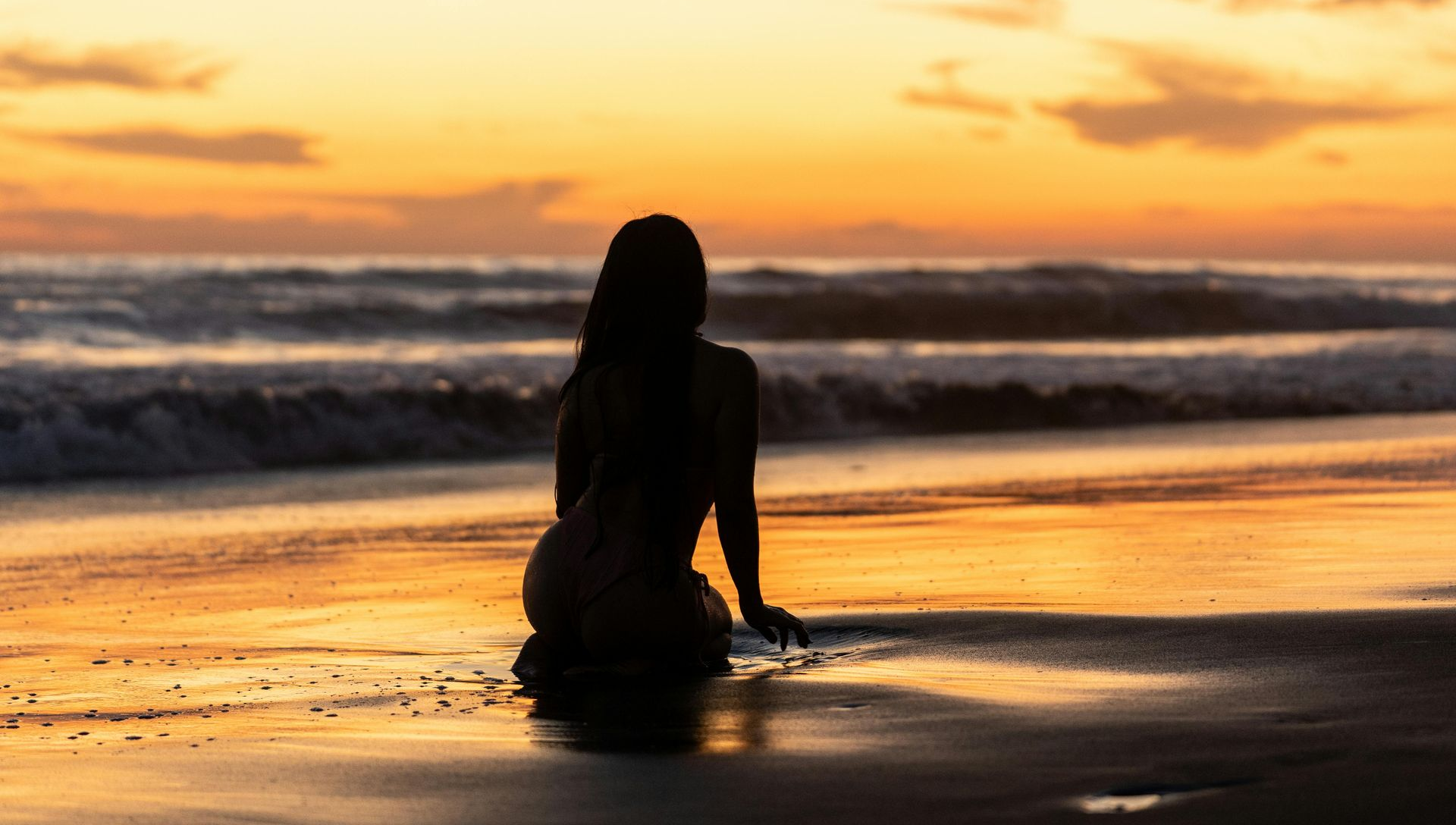 Silhouette of a person kneeling on a beach, watching the sunset over the ocean.