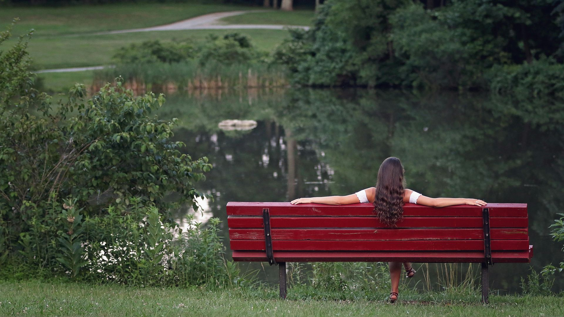 Woman sitting on a red park bench, arms outstretched, facing a pond and green foliage.