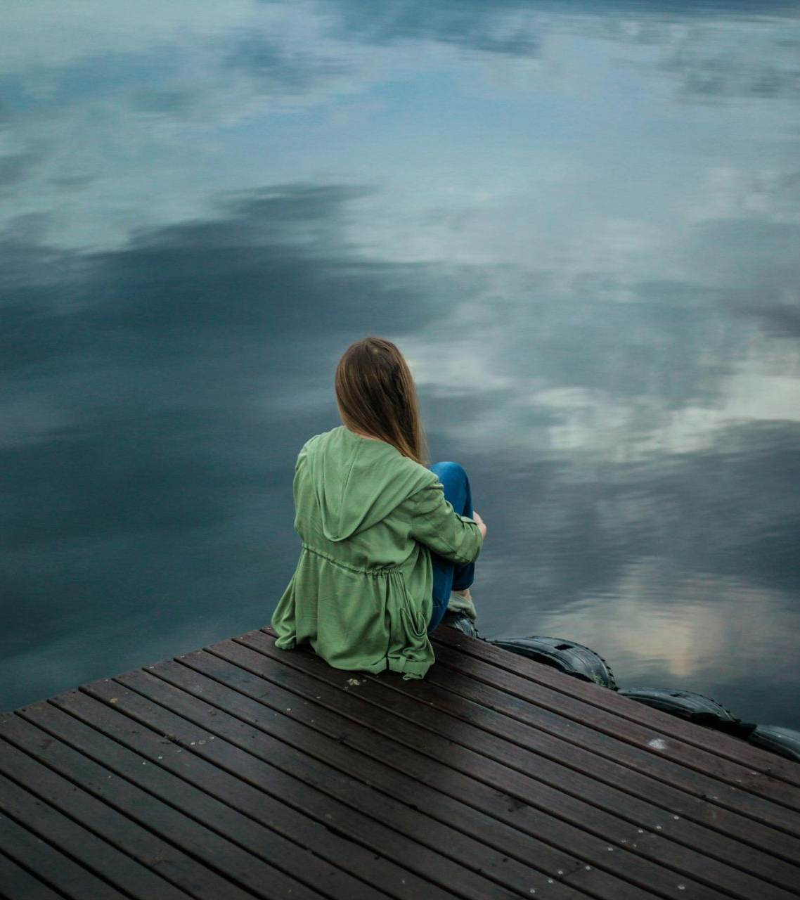 Woman sits alone on a wooden dock, hugging knees, overlooking water reflecting cloudy sky.