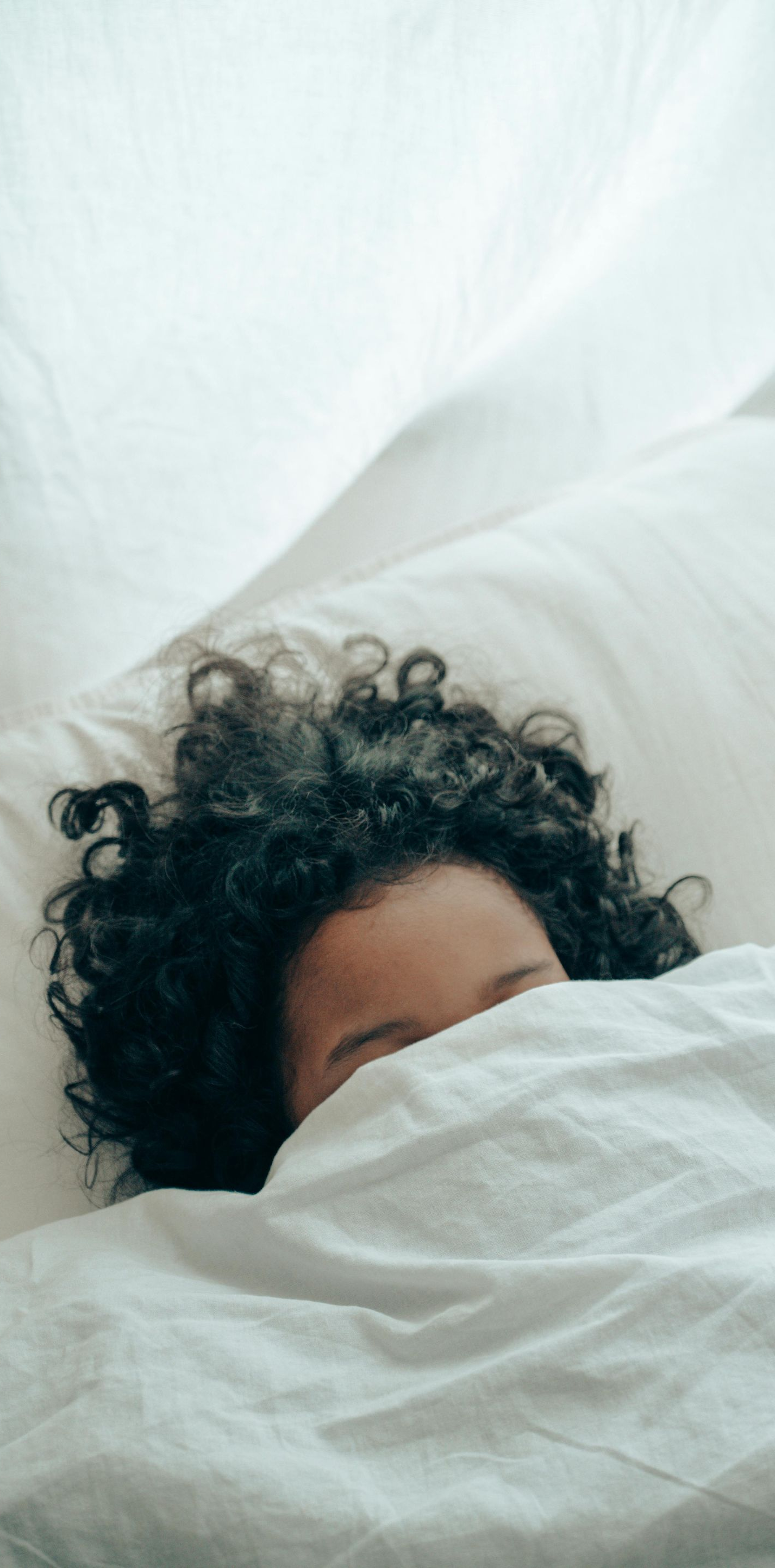 Person with curly hair, eyes hidden, under white sheets.