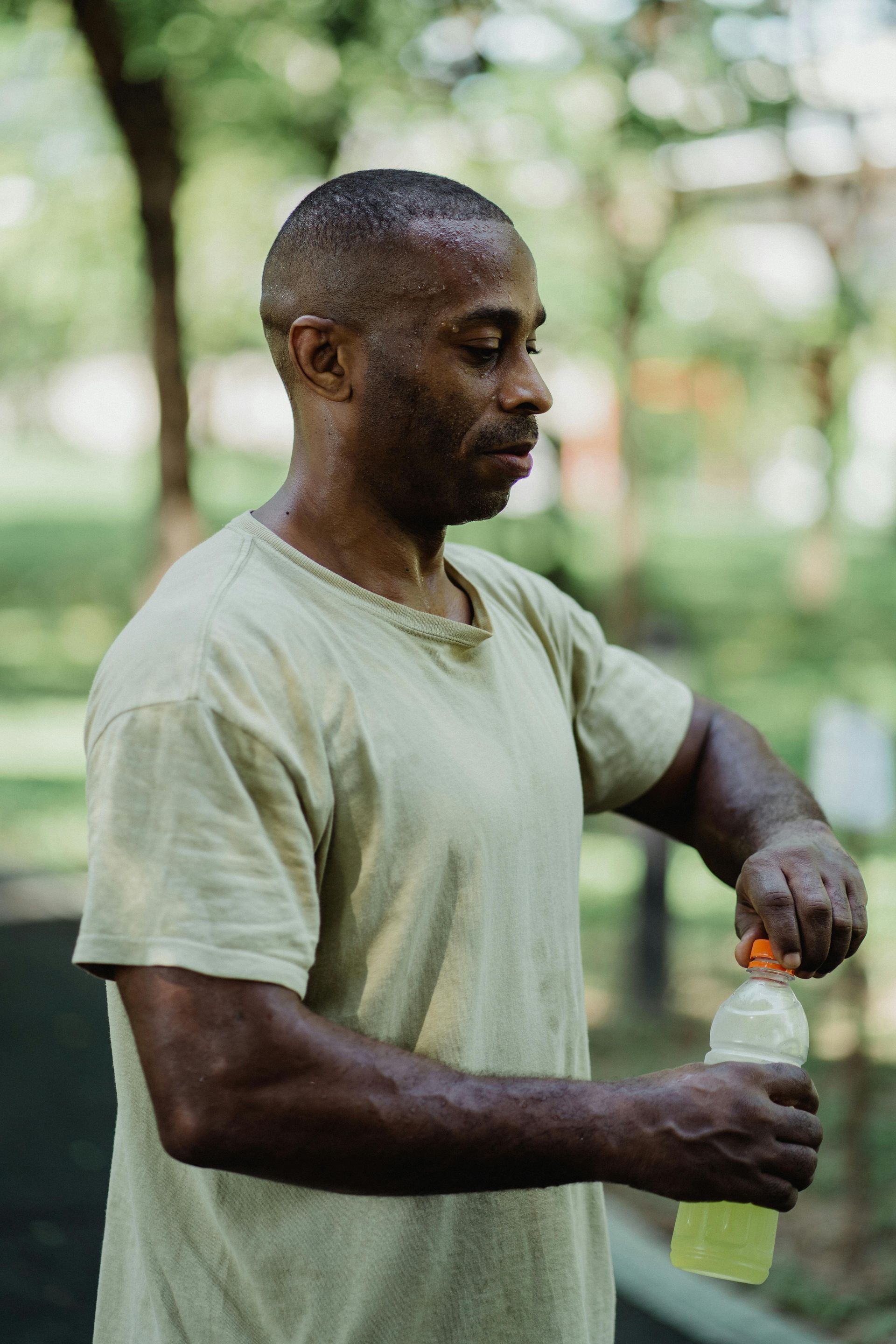 Man opening a sports drink bottle outdoors. He is sweating, in a light green shirt. Trees in the background.