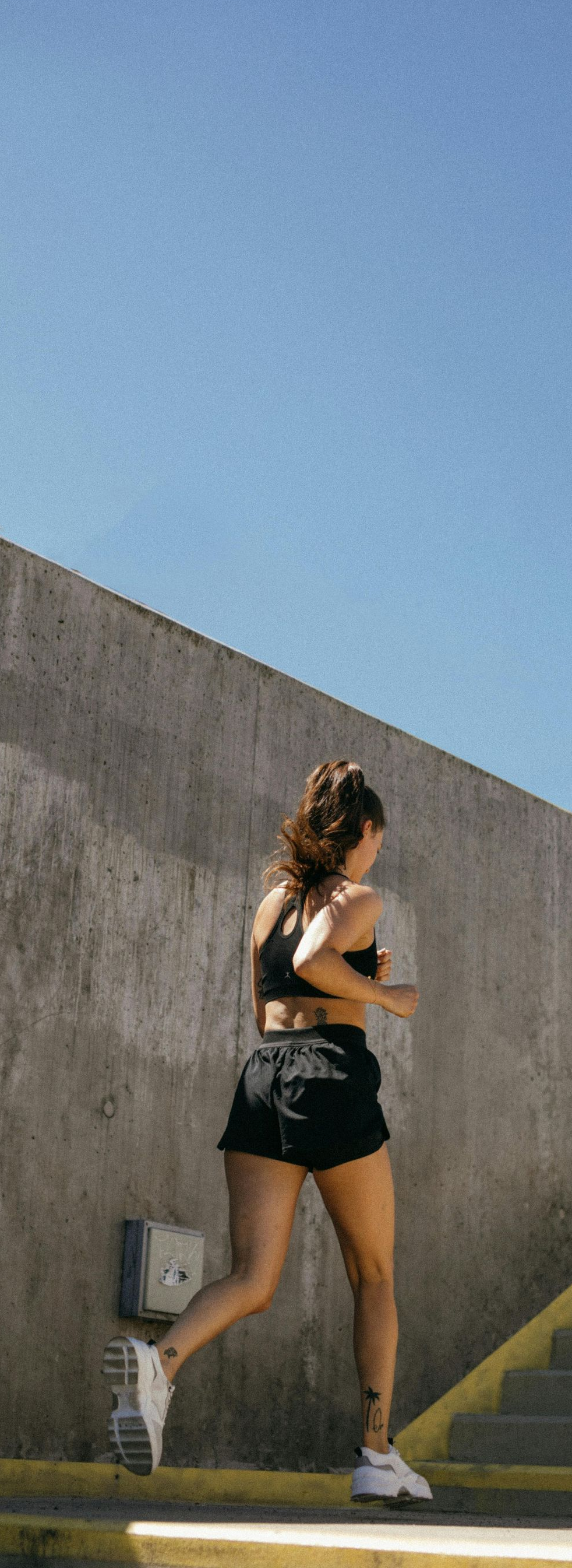 Woman running up stairs. She wears black shorts and top, white sneakers, outdoors against blue sky.