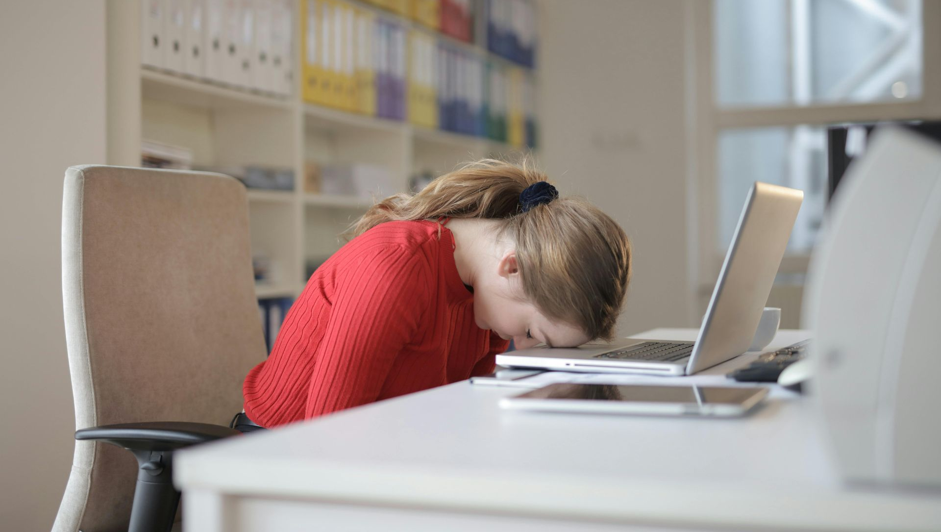 Woman in red shirt resting head on desk in front of a laptop and mobile devices.