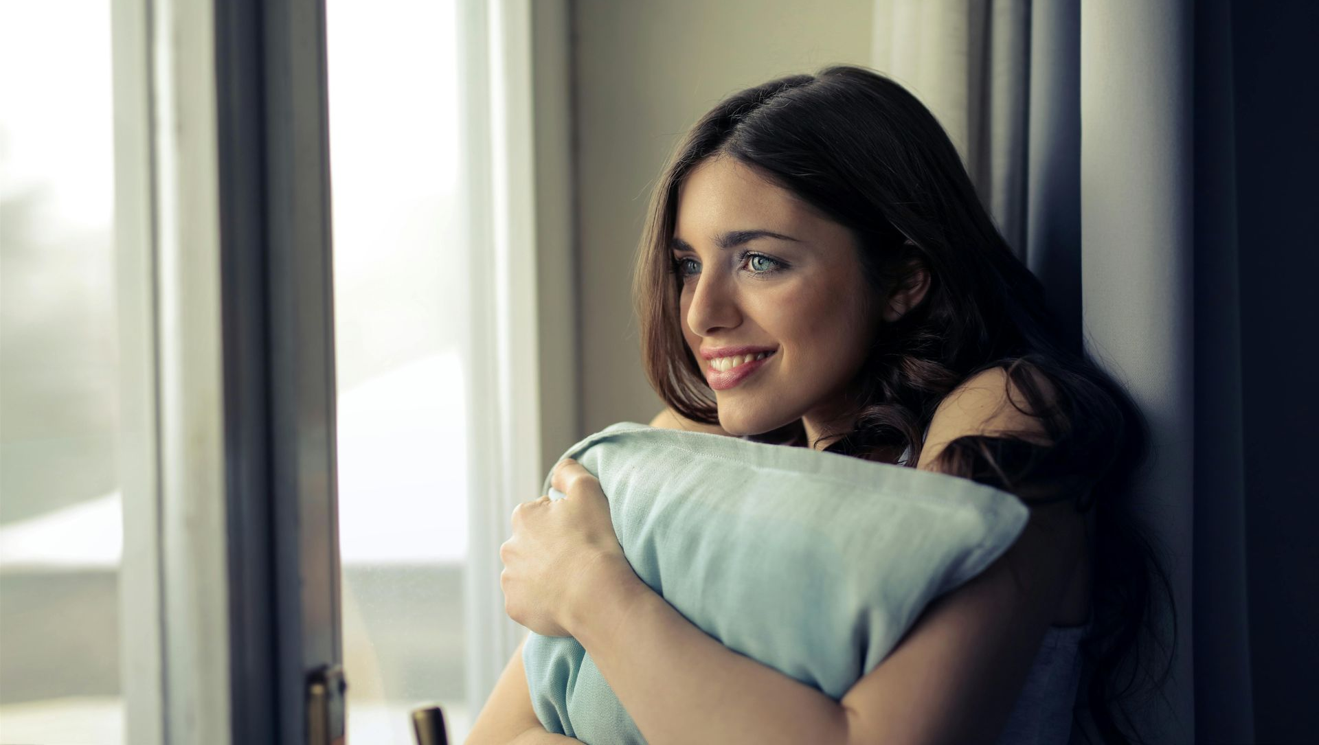 Woman smiling, hugging a light blue pillow, by a window.
