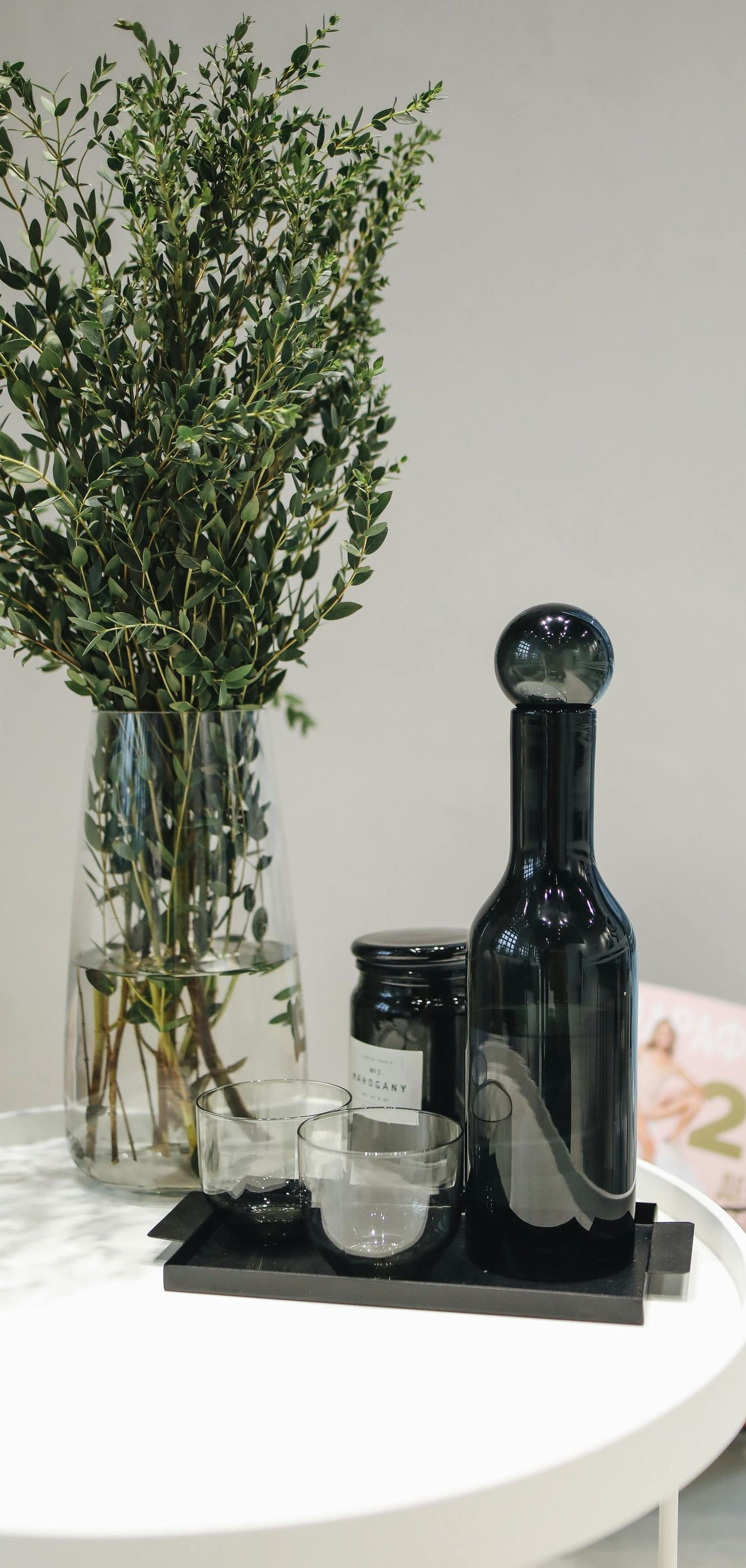 A glass vase with leafy greens, a dark glass bottle with a ball top, and other containers on a tray.