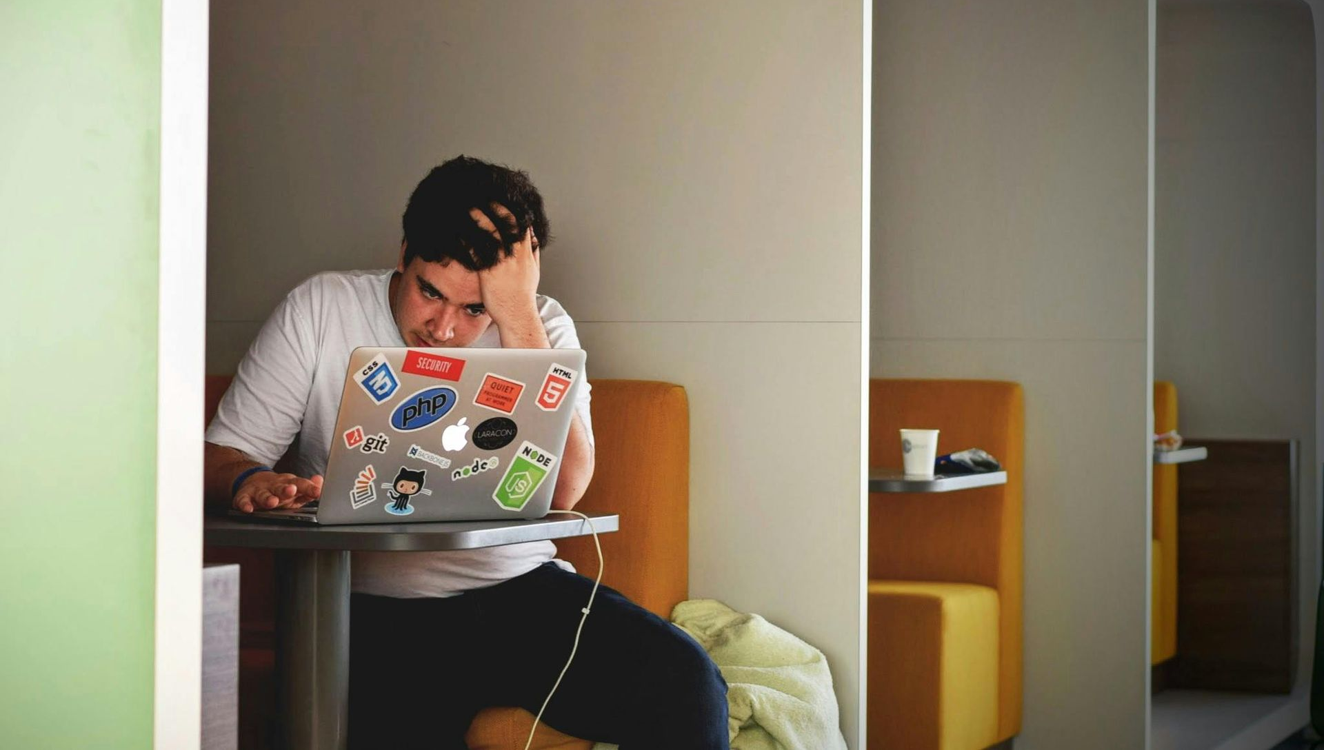 Person sits at desk, laptop open, hand on head, looking stressed. Coffee and light-colored space.