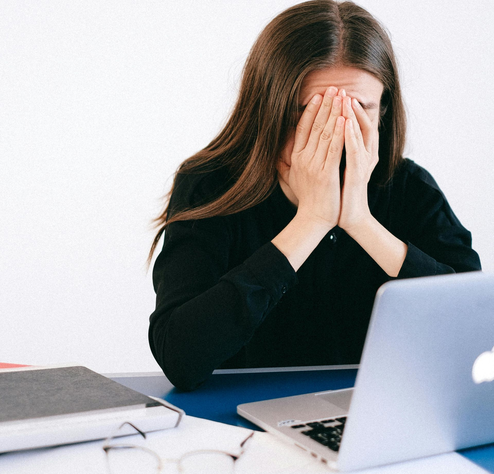 Woman with hands covering her face, sitting at a desk with a laptop, appearing stressed.