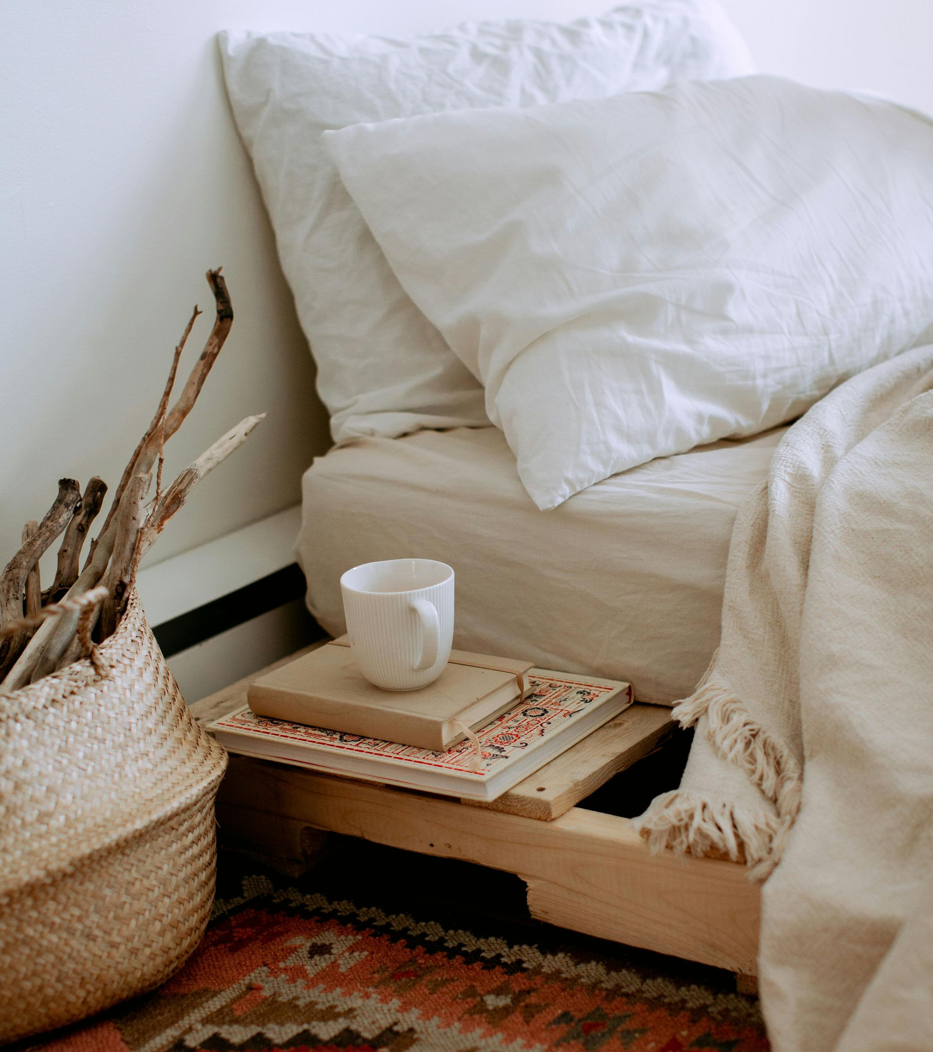 Bed with white linens, books, a mug, and a woven basket next to a colorful rug.
