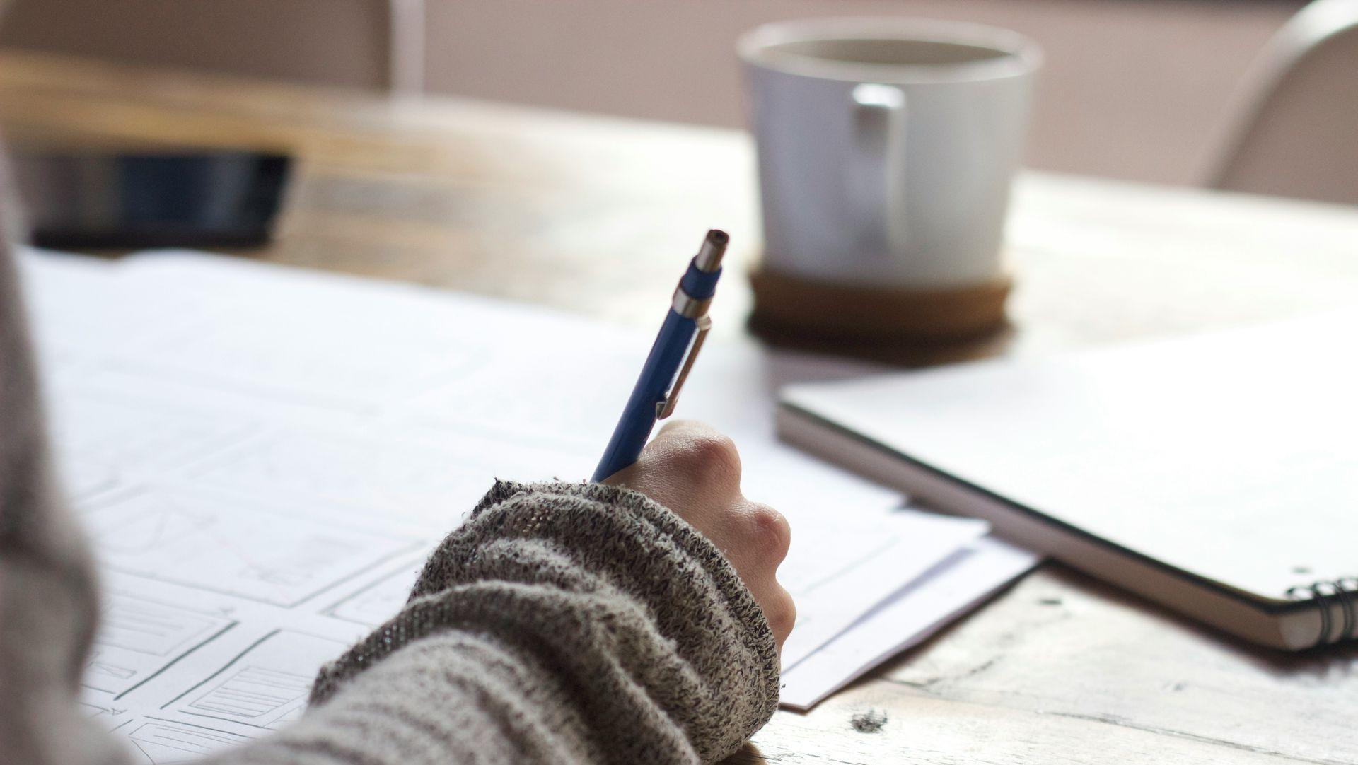 Person writing with a pen on paper, beside a mug and notebook on a wooden table.