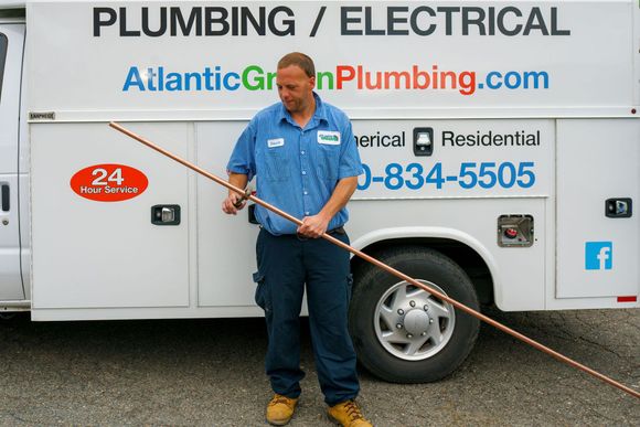 A plumber in a blue uniform holds a long copper pipe in front of a service van labeled AtlanticGreenPlumbing.com.