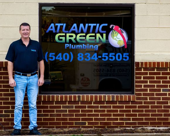 A person standing in front of a window featuring the Atlantic Green Plumbing logo and phone number.