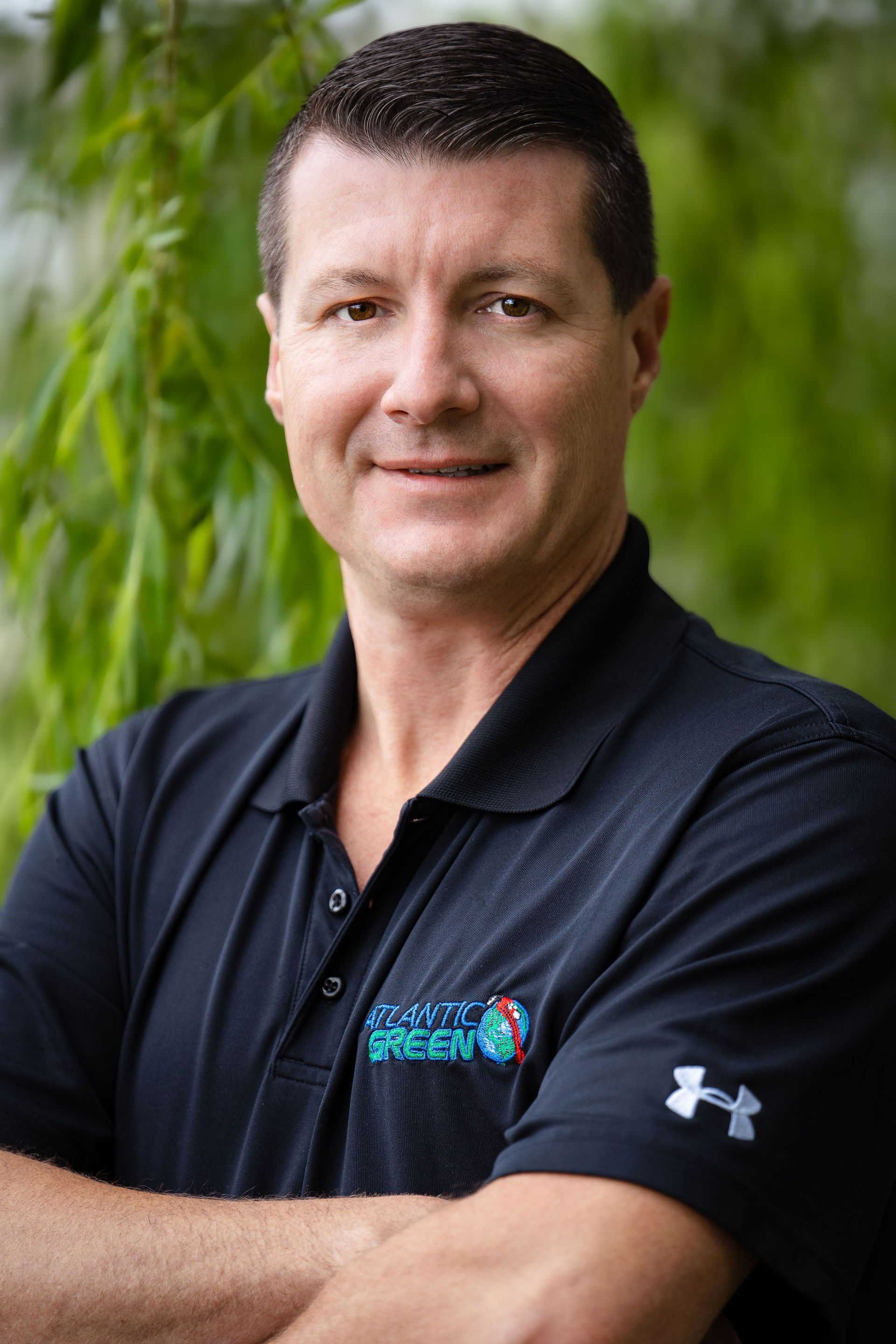 A smiling person with crossed arms, wearing a black polo shirt with a logo, standing in front of green foliage.