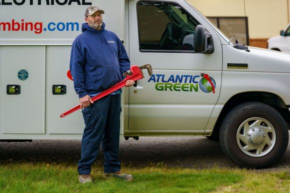 A worker holding a large red pipe wrench stands next to a white service van labeled 