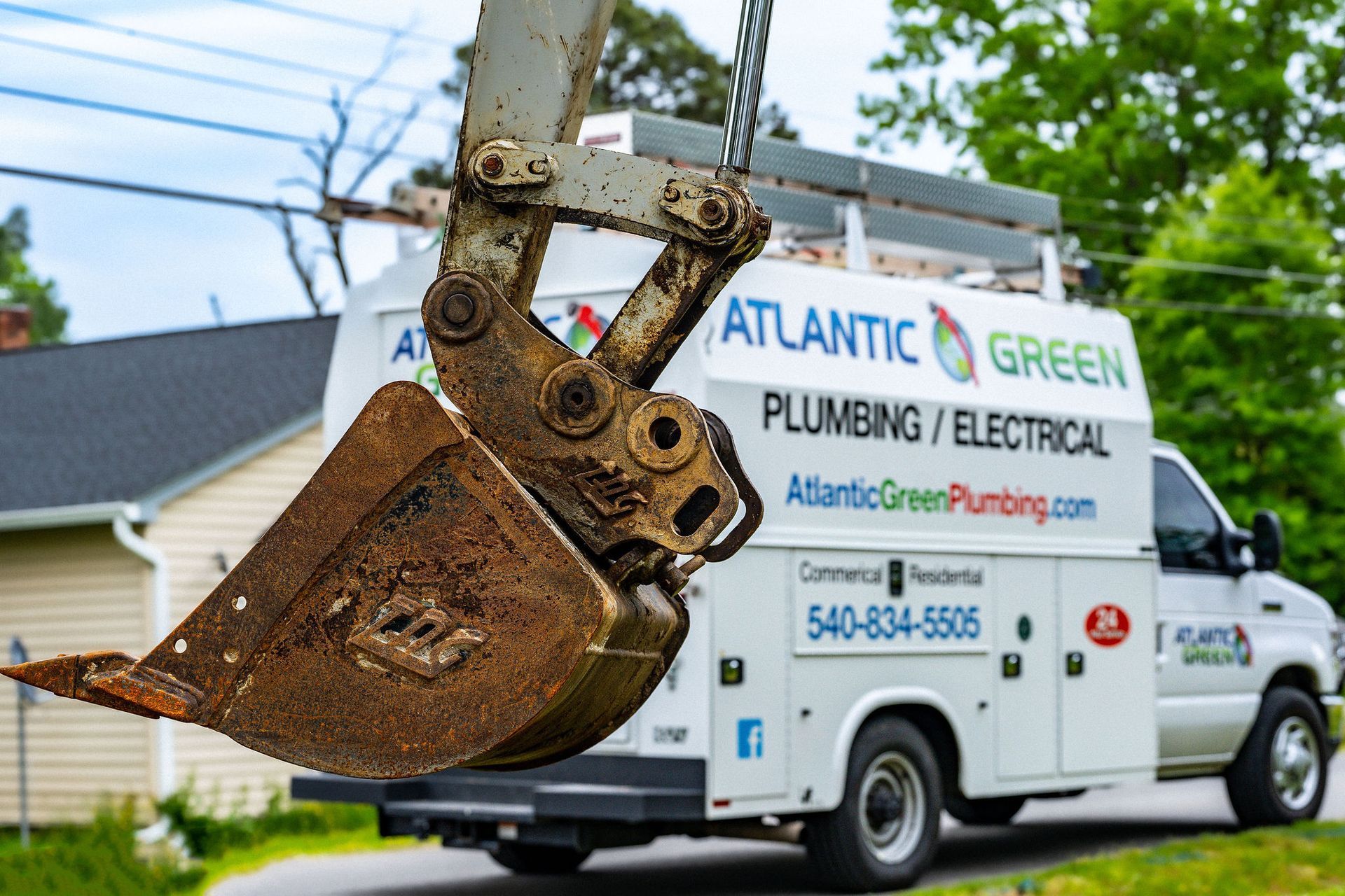 A muddy excavator bucket sits in the foreground with an Atlantic Green plumbing and electrical service van behind it.