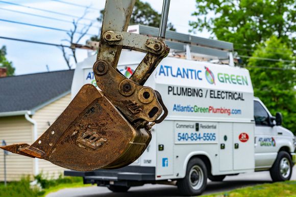 A muddy excavator bucket sits in the foreground with an Atlantic Green plumbing and electrical service van behind it.