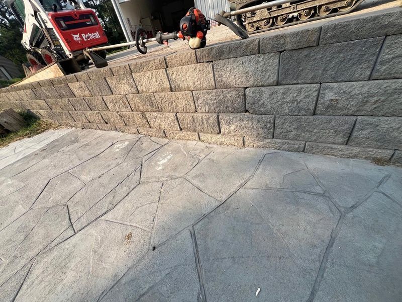 A low gray stone retaining wall separates a paved flagstone-style patio from a construction area with a red Bobcat loader.