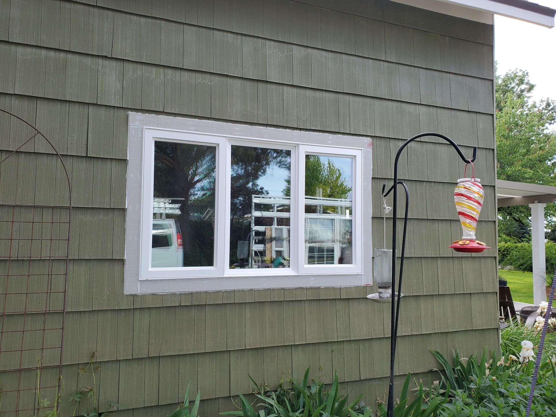Small white-trimmed window in a green shingled house exterior with a hanging bird feeder nearby.