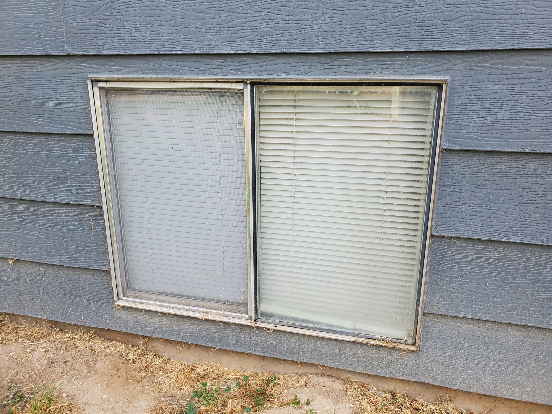 Two-pane window with blinds on a blue-gray house exterior and gravel below