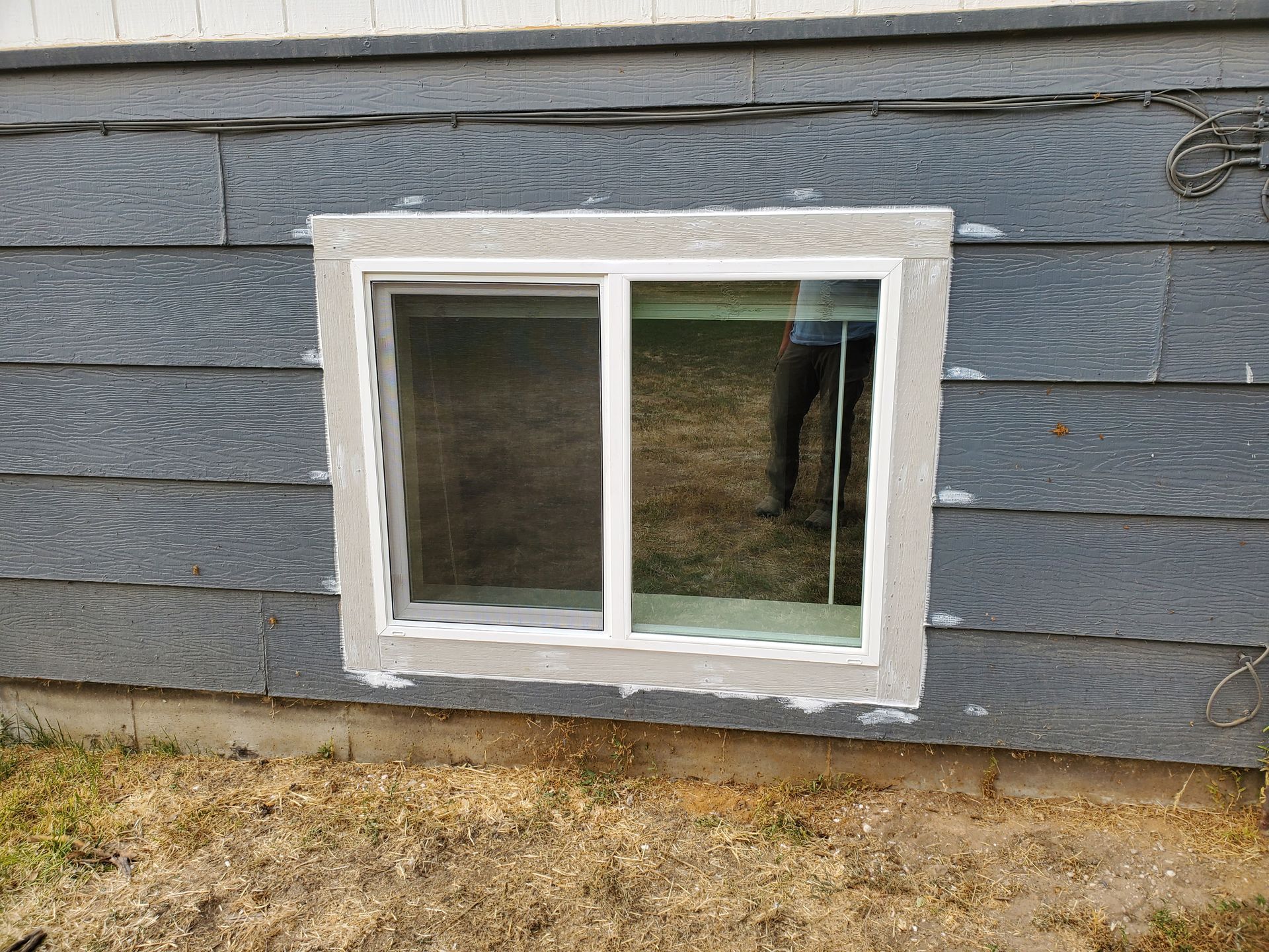 Small basement window with white trim in a gray wood-sided house exterior.