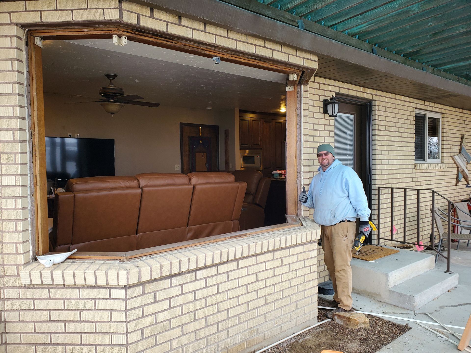 Man standing beside a damaged brick patio wall opening into a home interior with a brown sectional sofa.