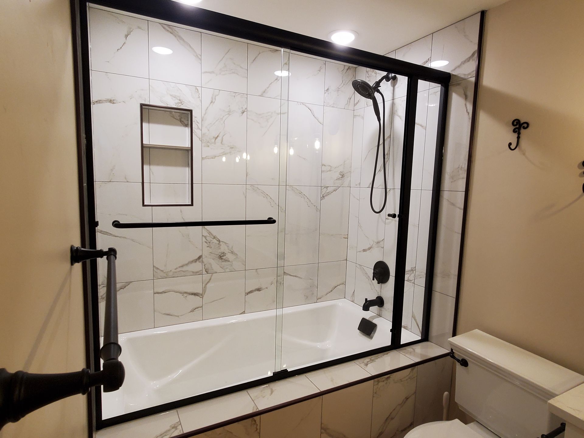 Modern bathroom with black-framed glass shower enclosure, marble tile walls, and a bathtub inside.