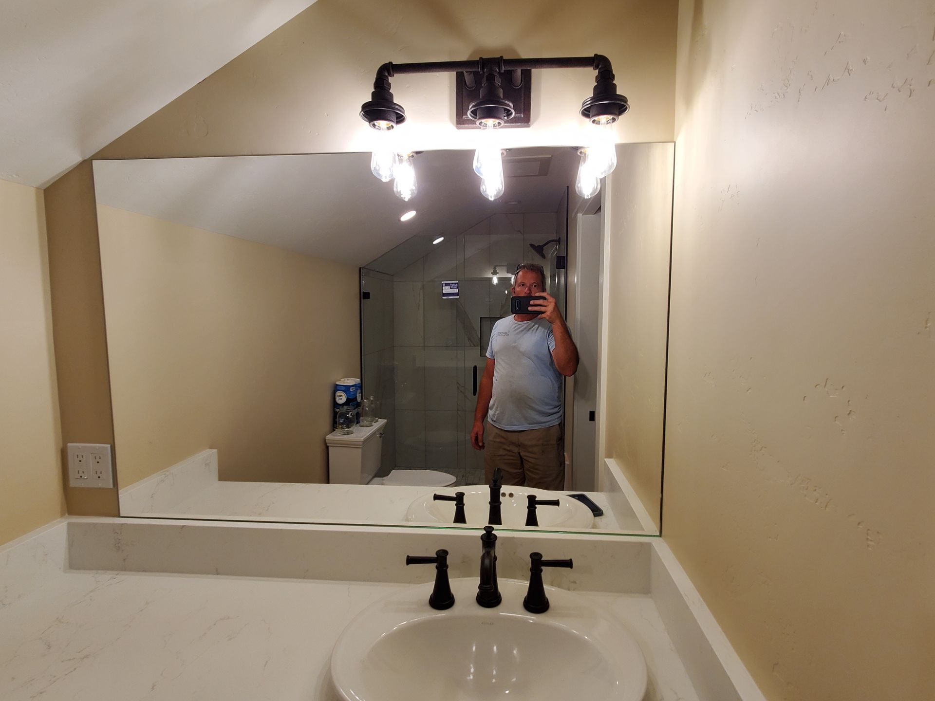 Bathroom mirror selfie in a beige restroom with a sink and bright overhead lights.