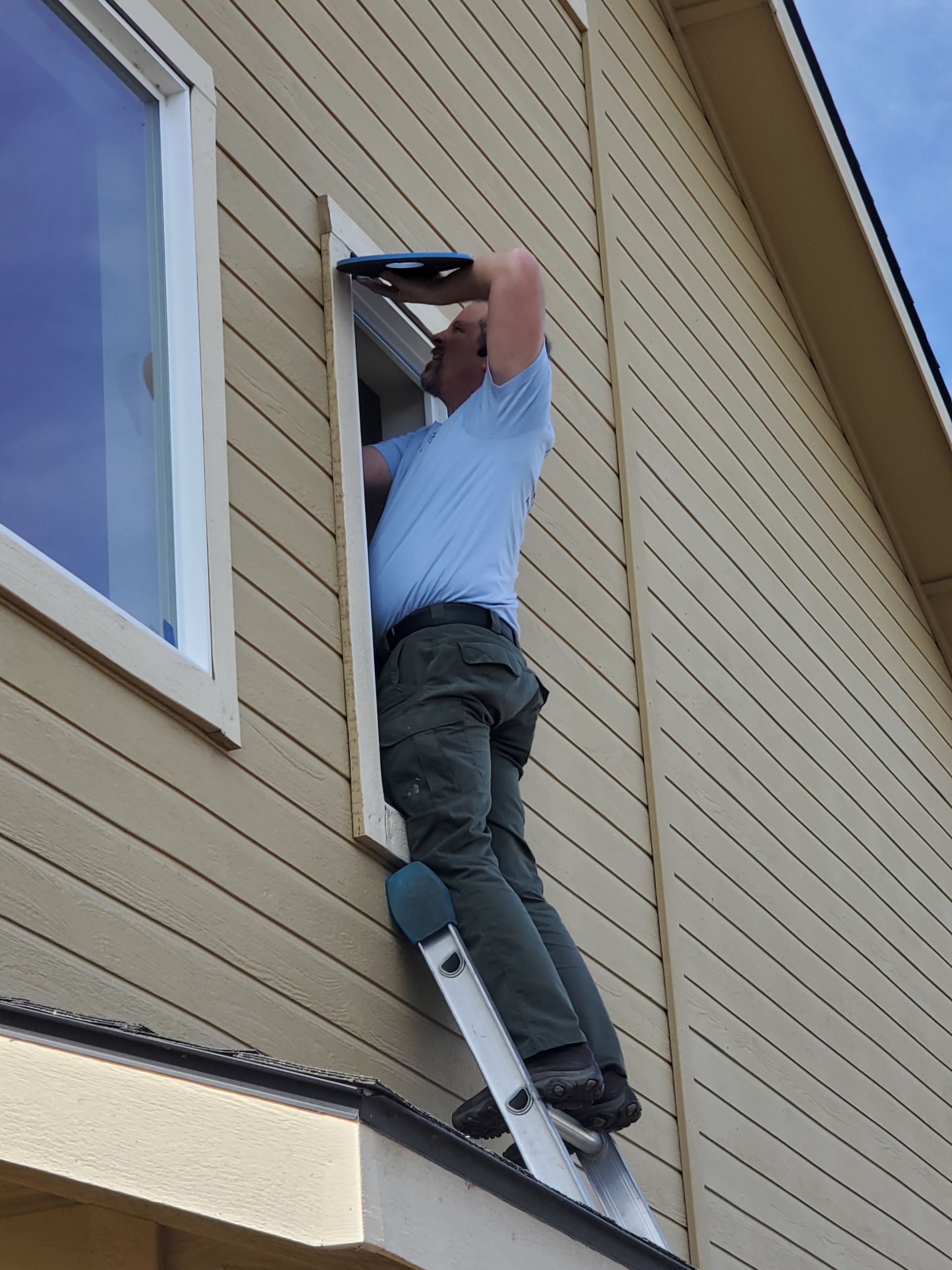 Person on a ladder working on the exterior of a beige house near a window.