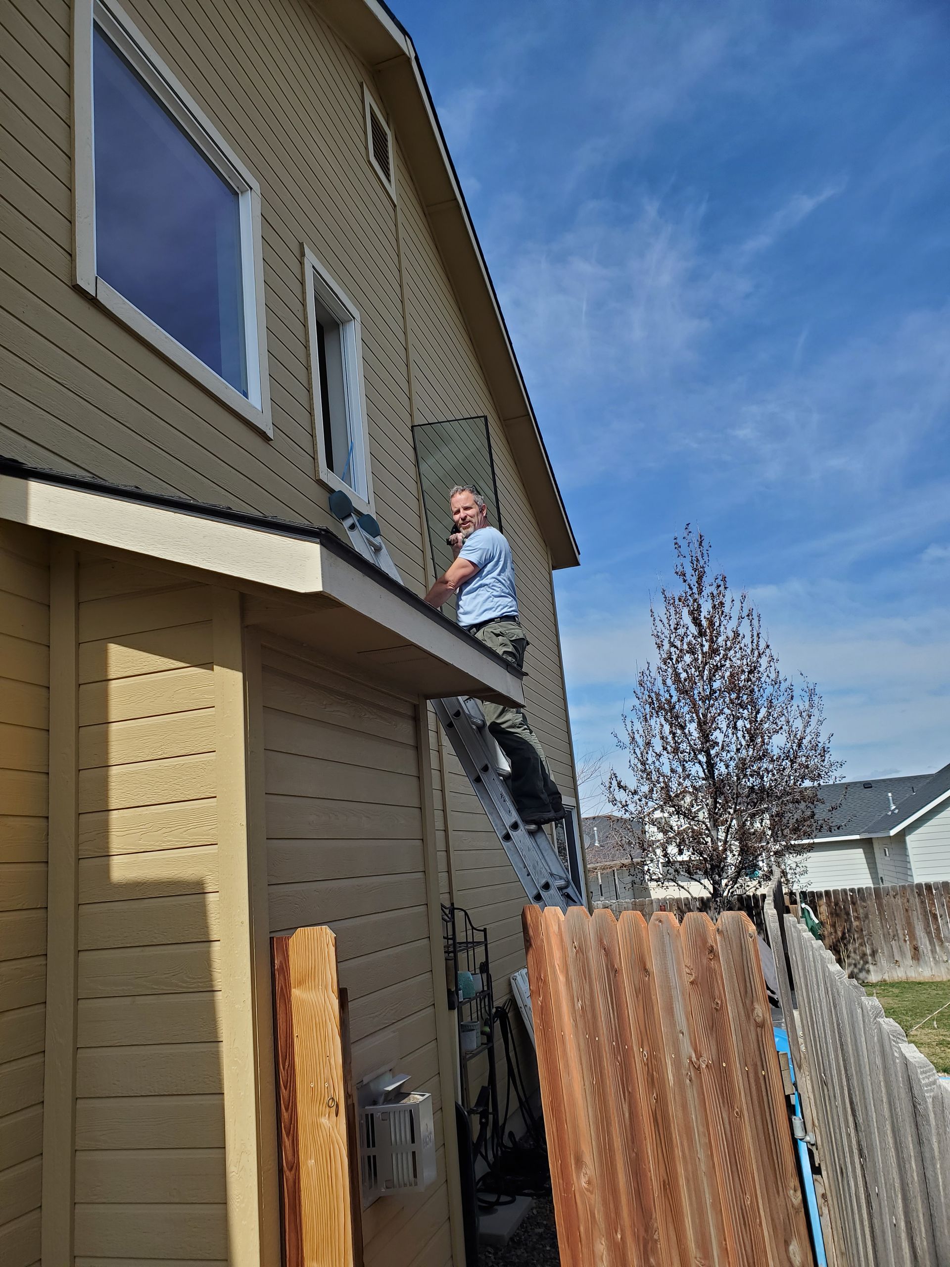 Person standing on a ladder beside a tan house with a wooden fence under a blue sky