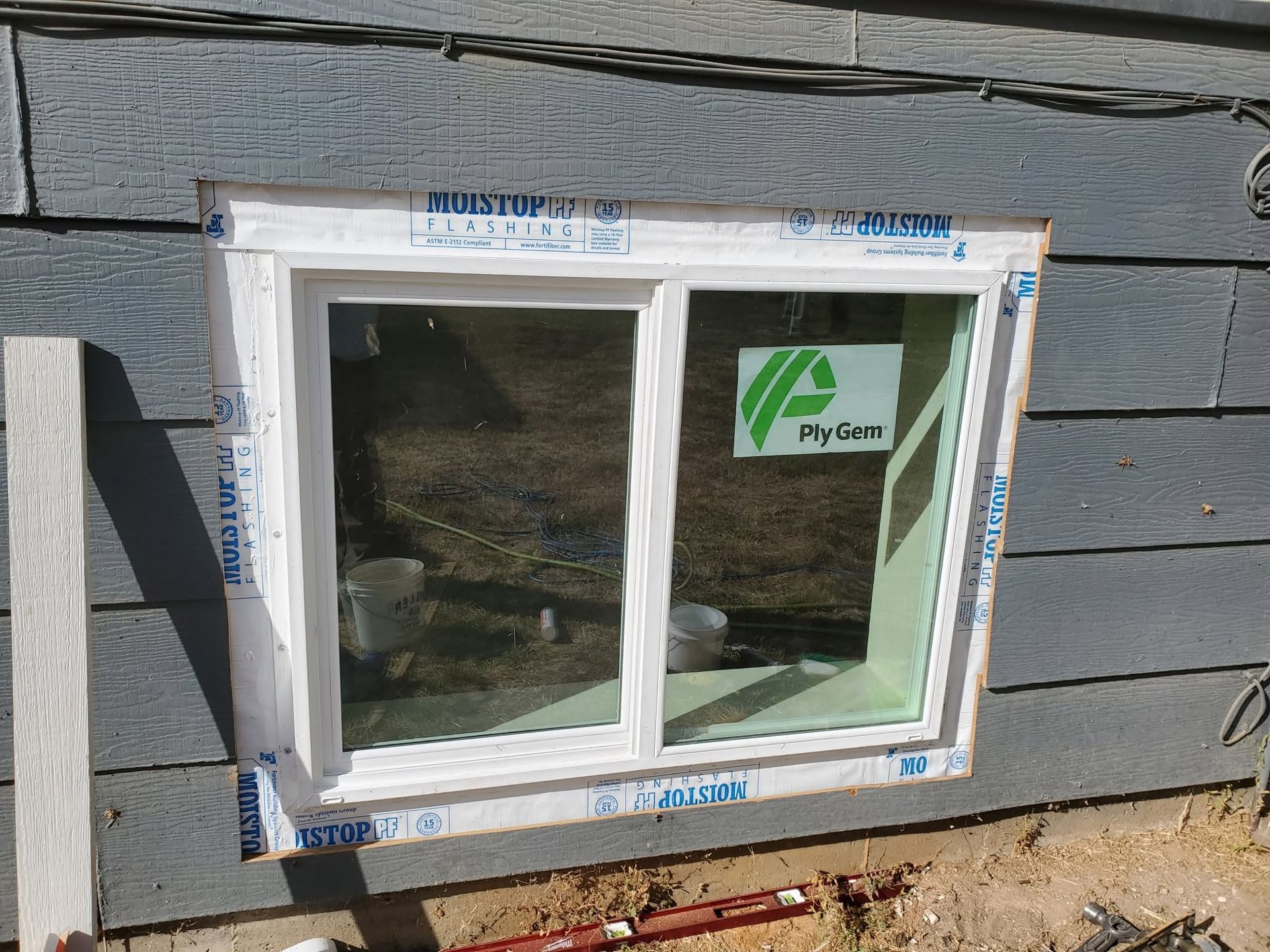 Small white-framed sliding window set into gray shingle siding with blue masking tape around it.