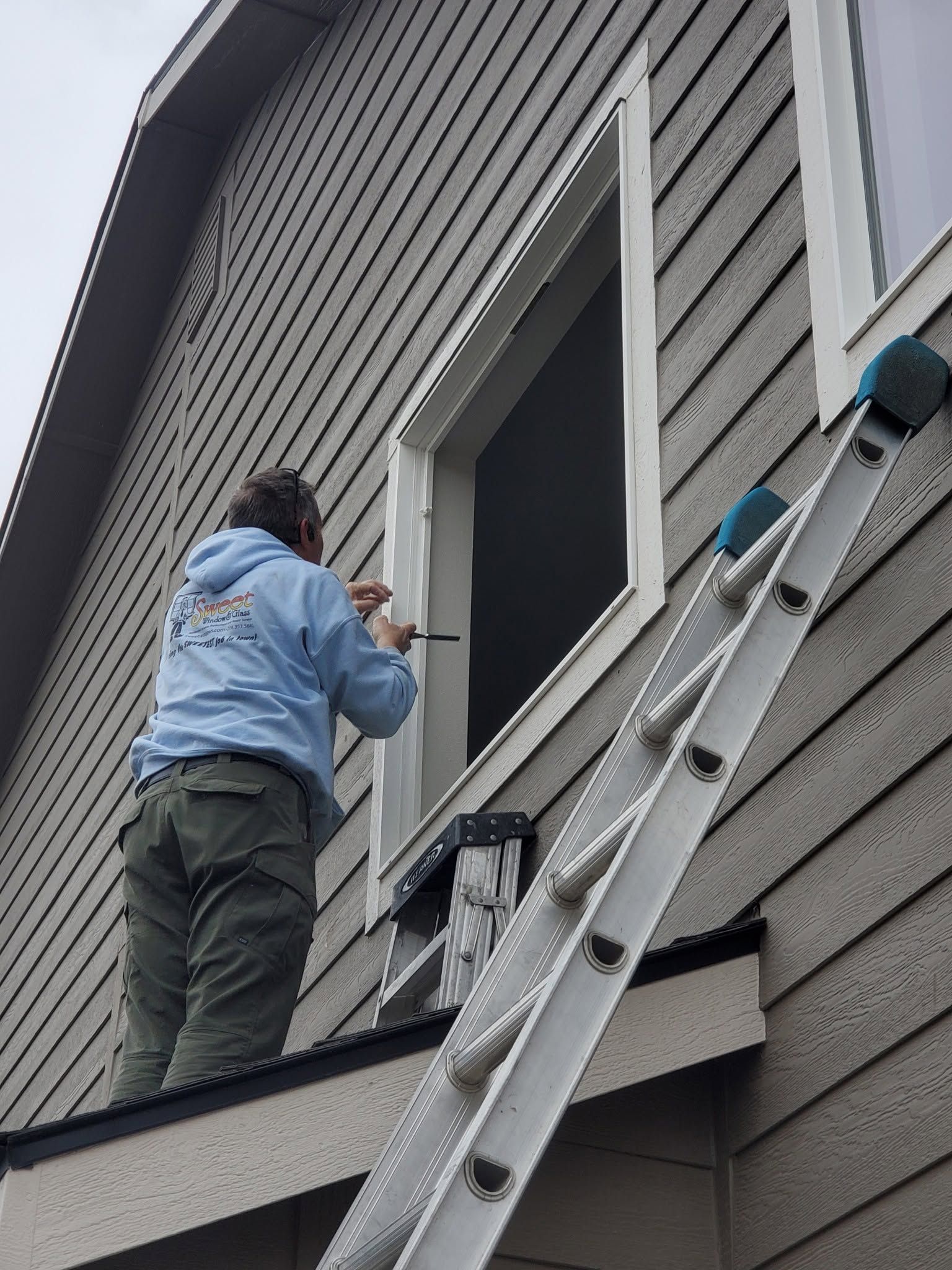 Person working on house siding beside an open window, standing on a ladder with tools.