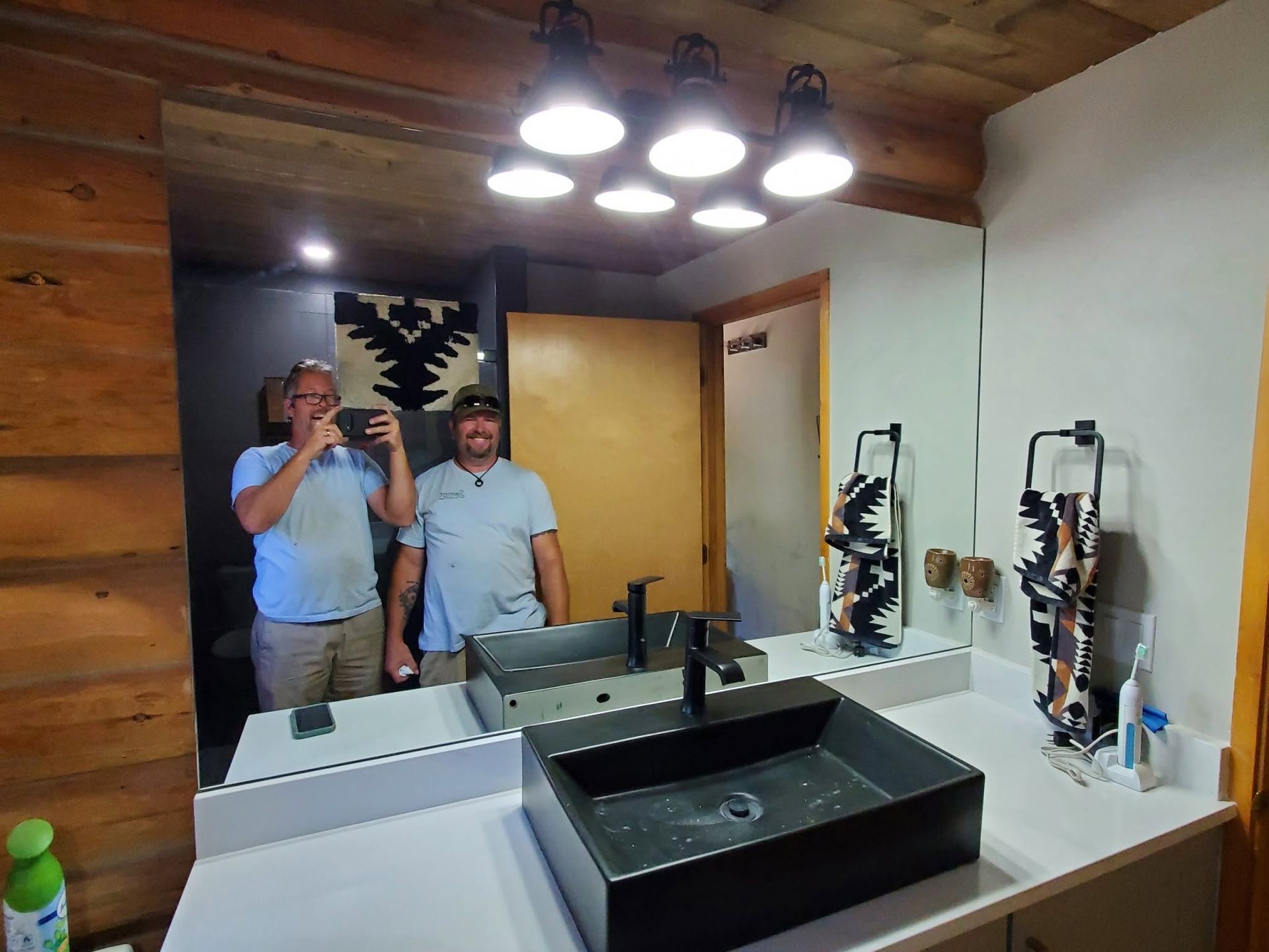 Bathroom vanity mirror showing a person taking a selfie under bright overhead lights.