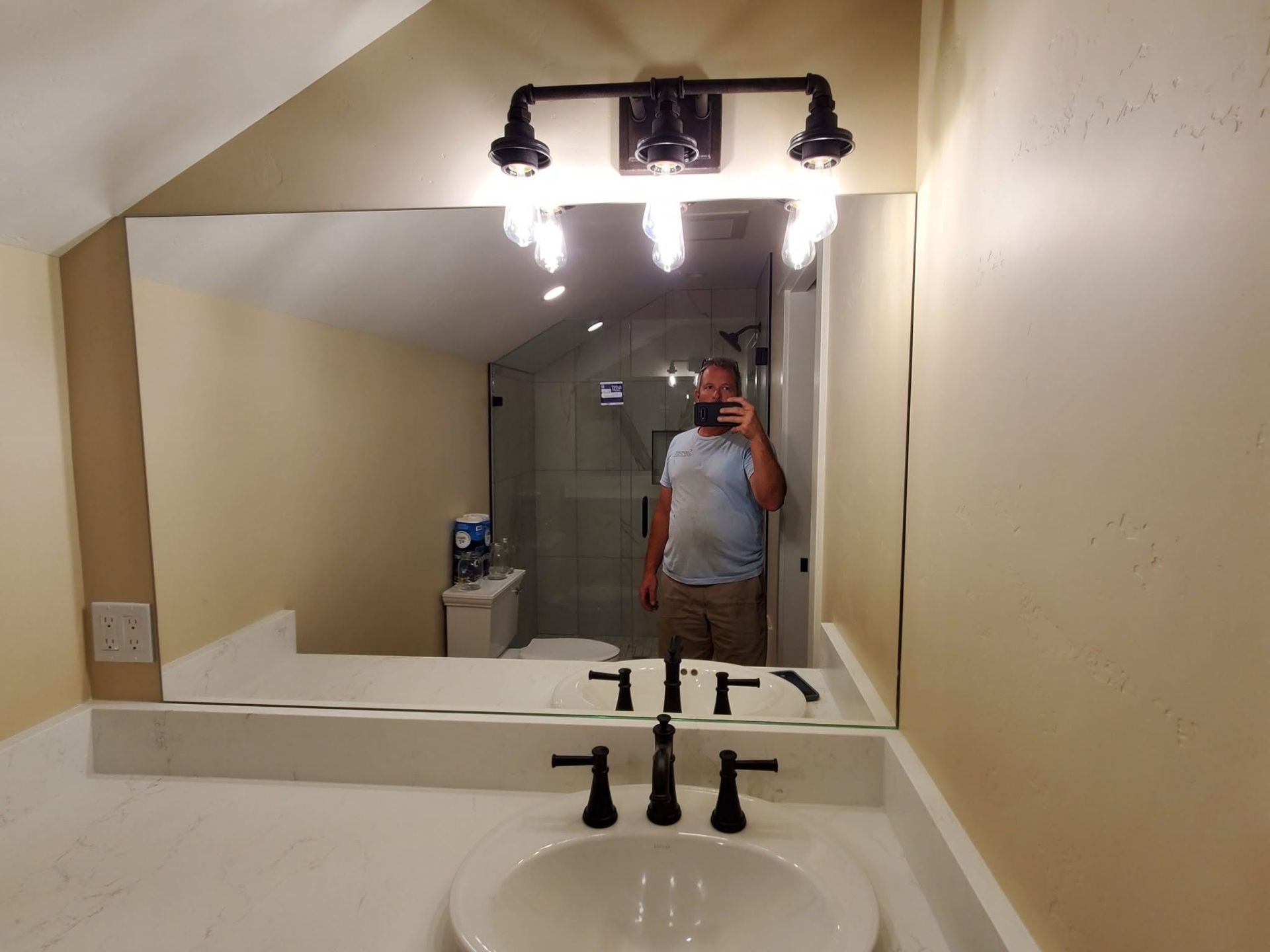 Bathroom mirror reflecting a person taking a selfie at a sink with bright wall lights.