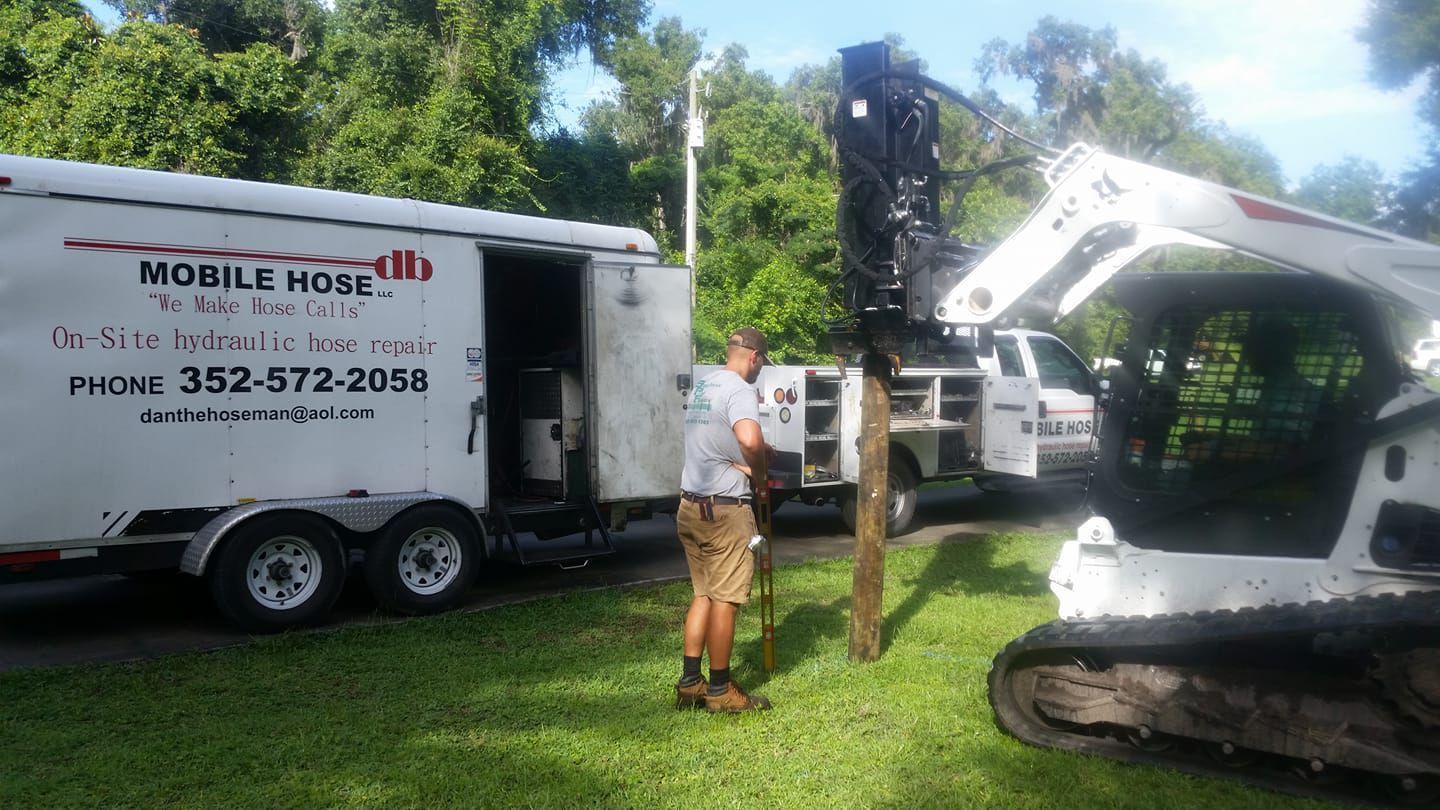 Man stands near a trailer and equipment, working on a utility pole in a yard.