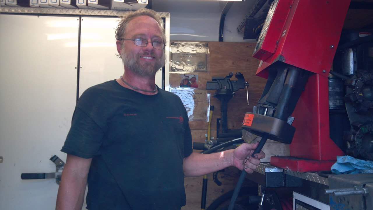 Man in a garage smiles, holding a tool; red machine and tools in the background.