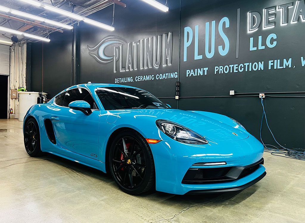 Bright blue Porsche coupe in a detailing shop with “PLUS DETAIL” wall branding