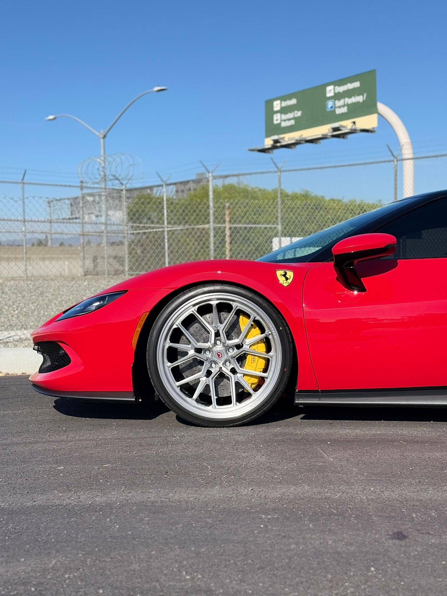 Red sports car parked on asphalt, front wheel in focus, with highway signs and fencing behind.