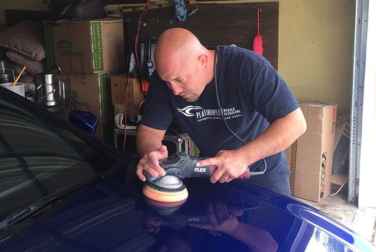 Man polishing a blue car hood with a buffer in a garage