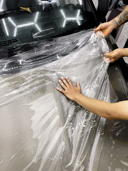 Hands peeling clear protective film from a glossy black car hood in a garage.