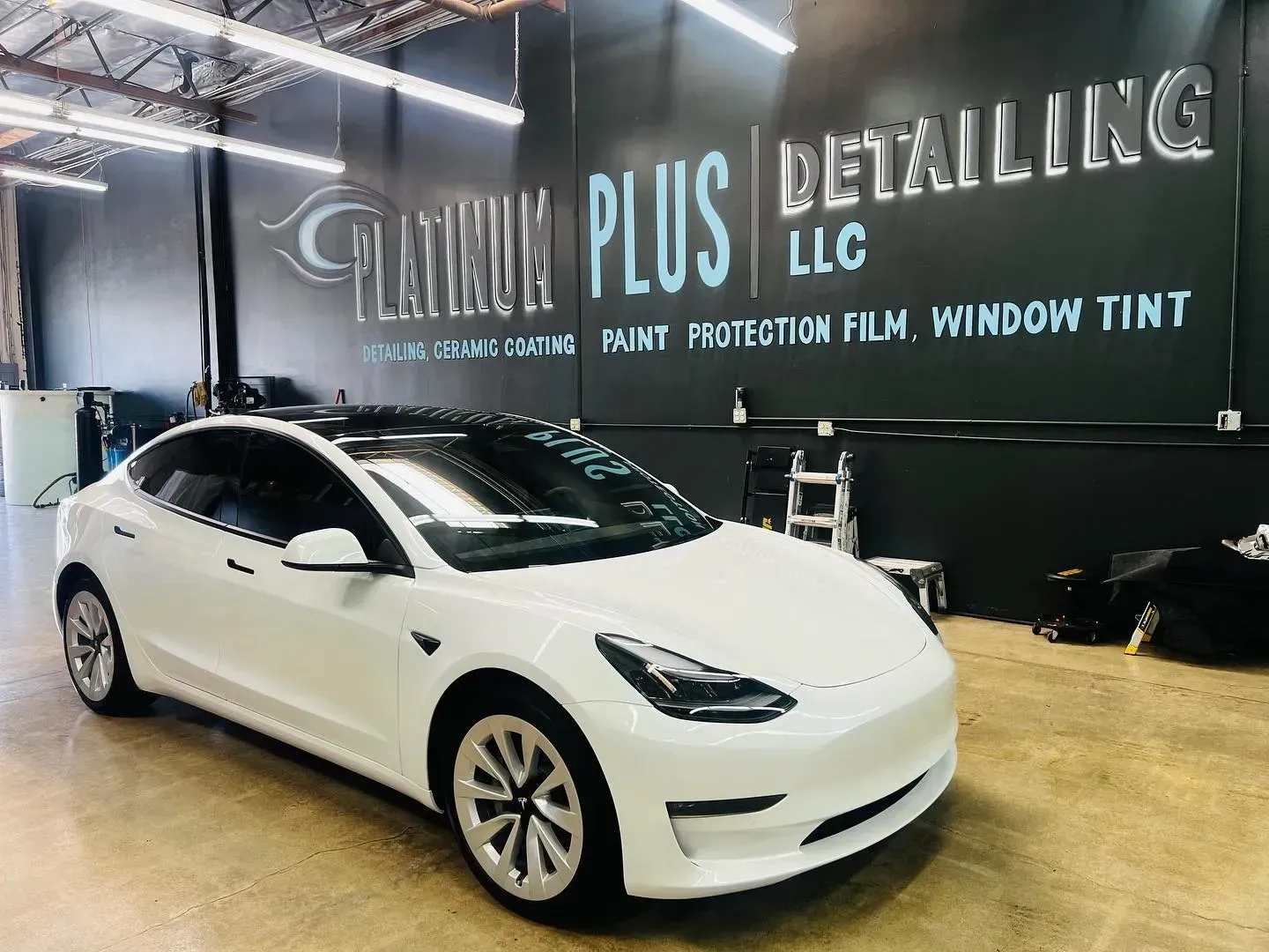 White Tesla sedan in a showroom with a “Detailing LLC” sign on the wall