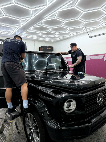 Two men polishing a black Mercedes G-Class in a bright studio with hexagon ceiling lights