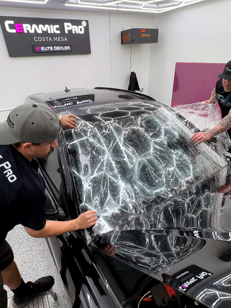 Two people applying ceramic coating to a black car in a garage, with a foam-covered windshield.