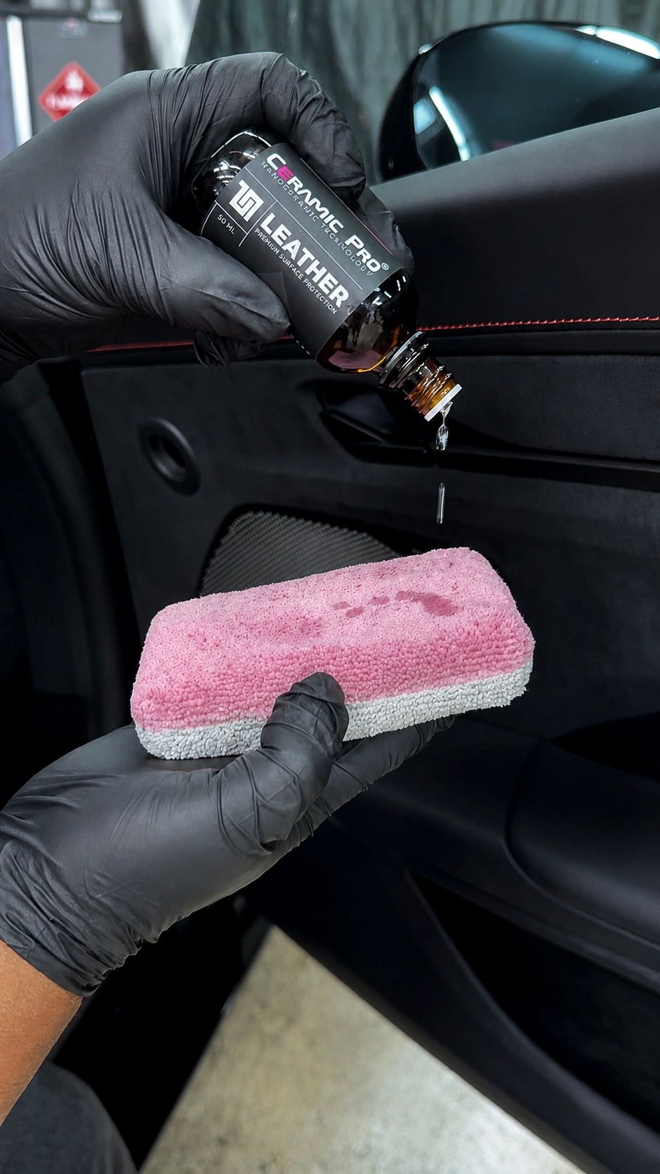 Gloved hands apply black cleaner to a pink applicator on a black car door panel.