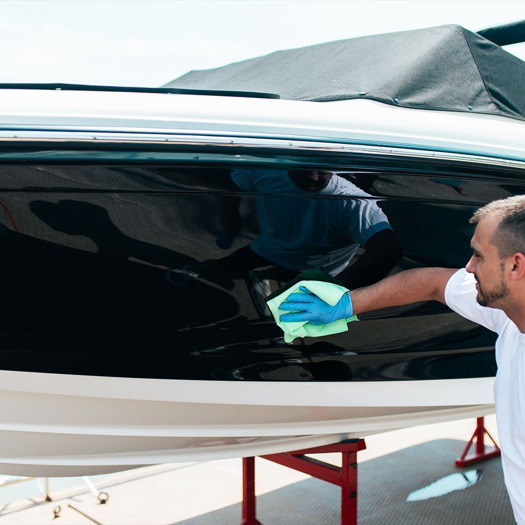 Person cleaning a shiny boat hull with a blue cloth on a trailer in bright sunlight