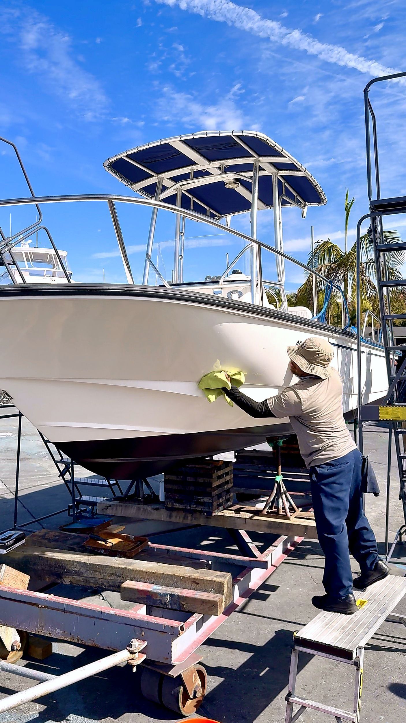 Man in beige shirt working on a white boat on a trailer beside a dock, under a blue sky