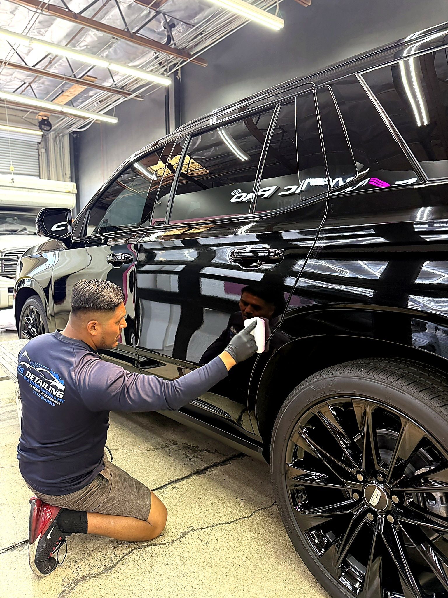 Man kneeling beside a black SUV, wiping the door in a garage under bright lights.