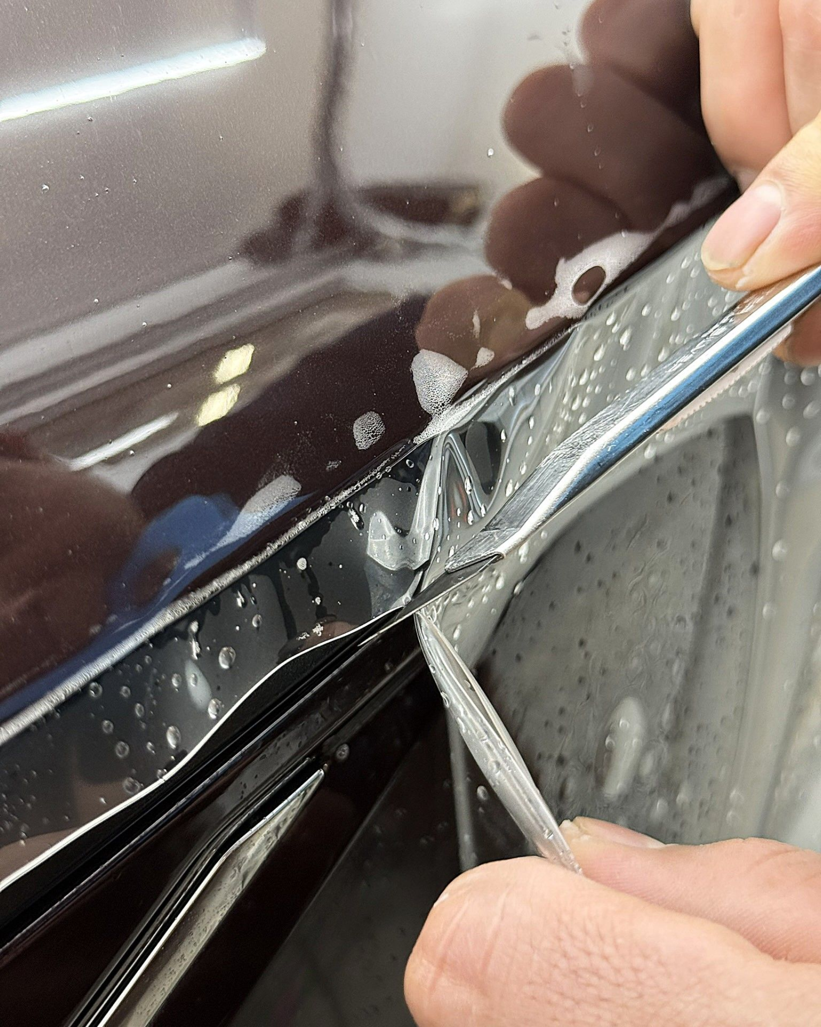 Hands peeling back black car trim with a metal tool near a glossy dark vehicle surface