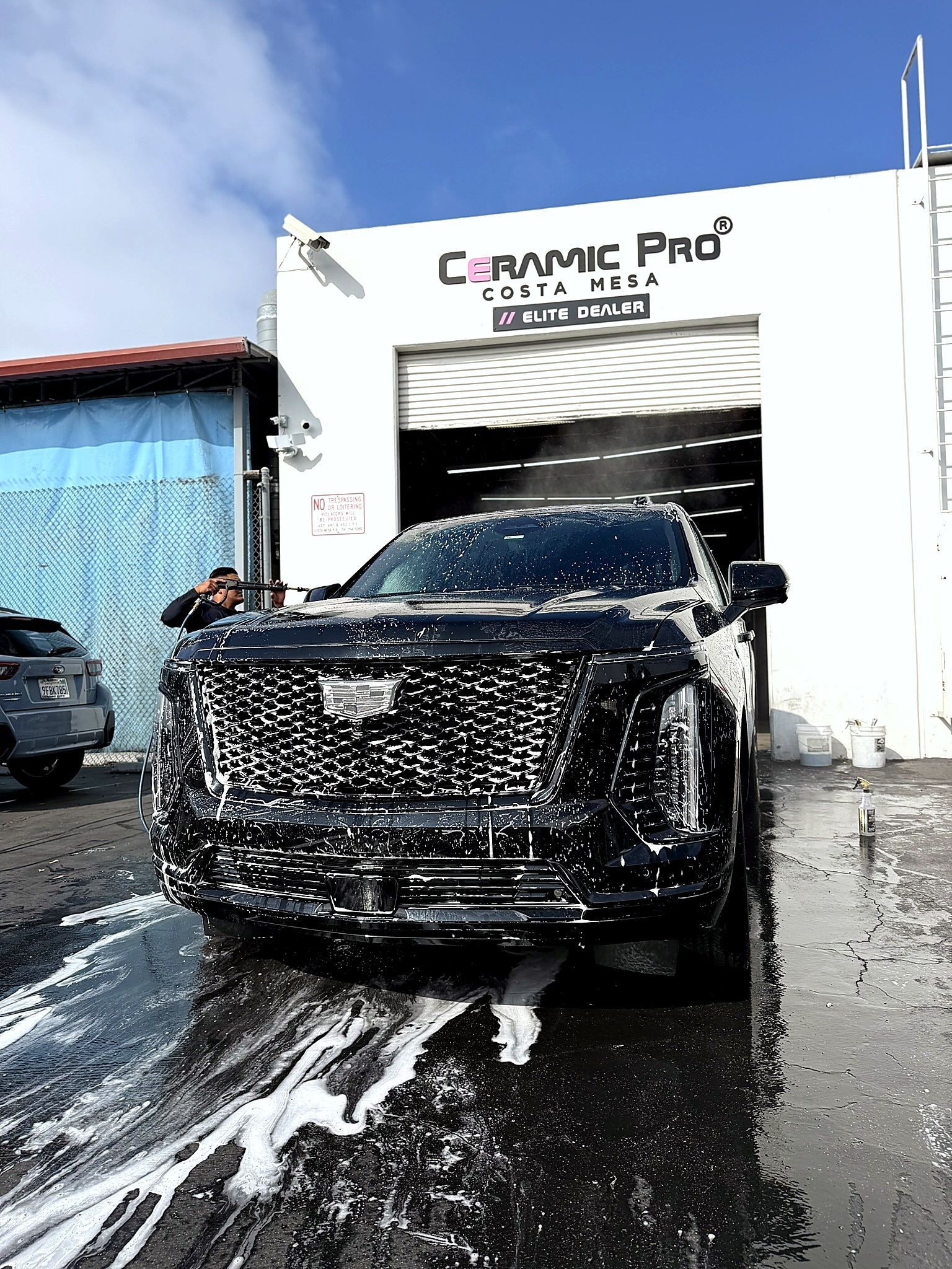 Black SUV being washed outside a car detailing shop, with soapy water on the ground.