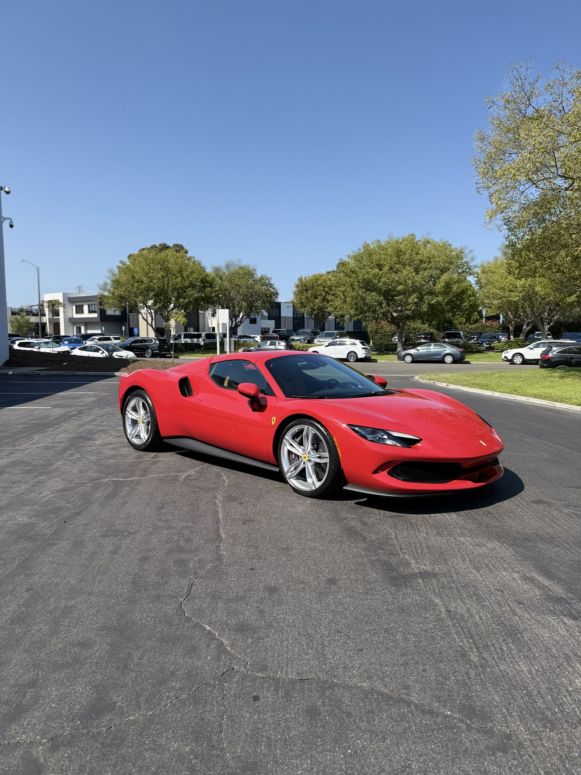Red sports car parked on a paved lot under a clear blue sky with trees and other cars in the background