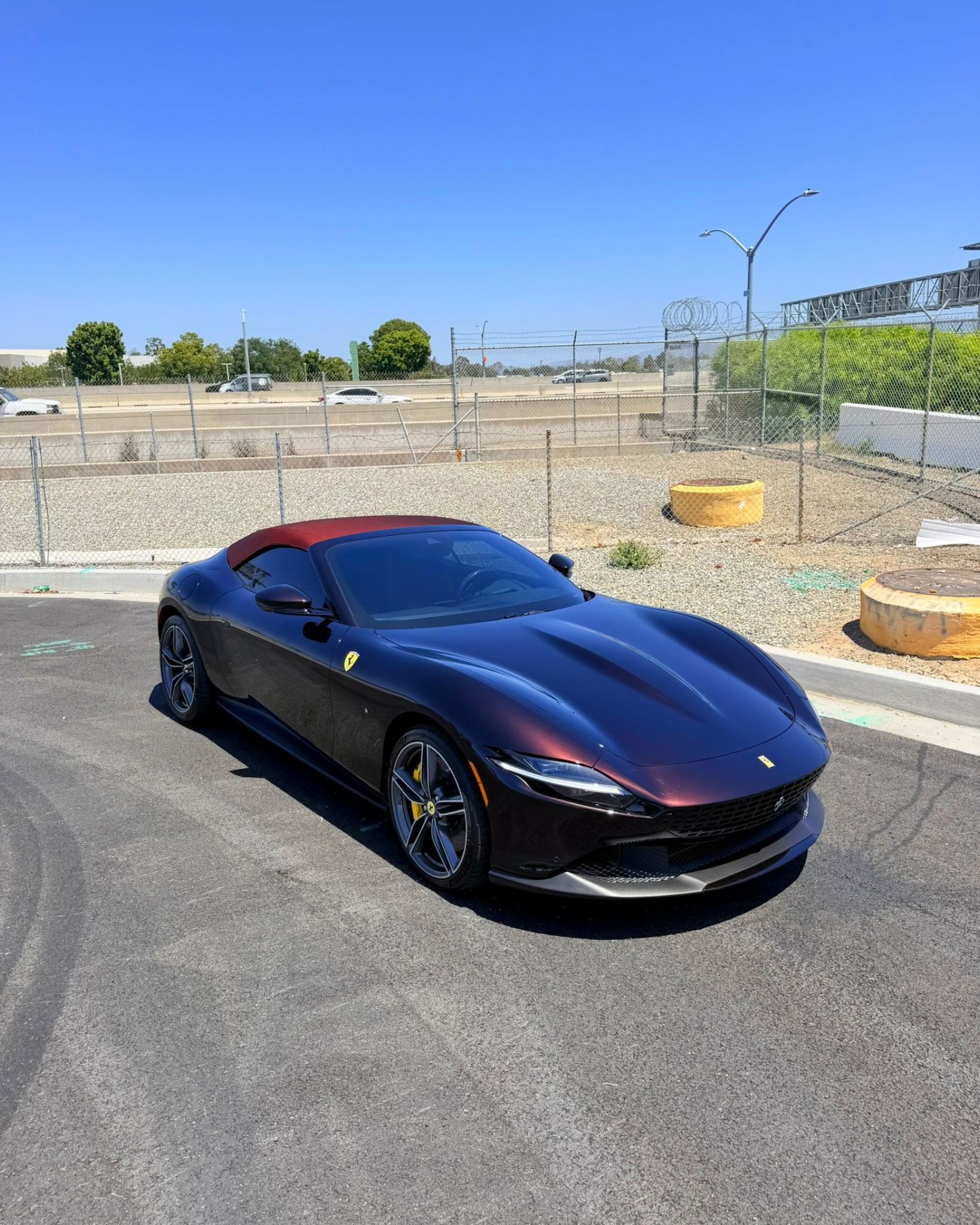 Black sports car parked on a sunny street beside a chain-link fence and construction area